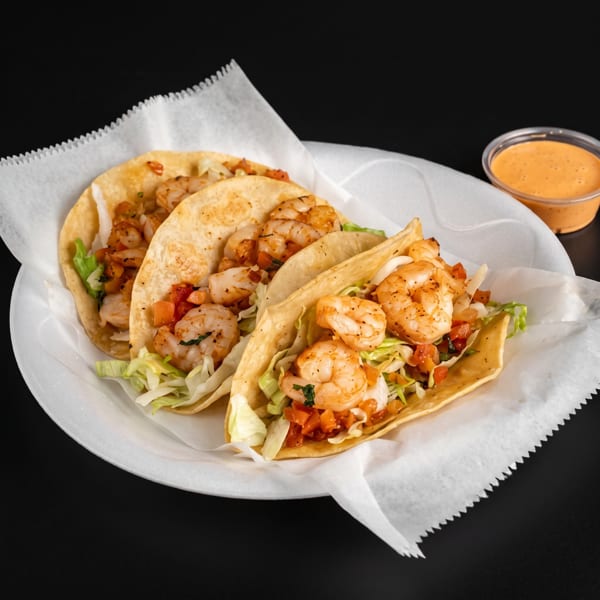 Three shrimp tacos with lettuce and diced tomatoes served on paper-lined plate with orange dipping sauce.