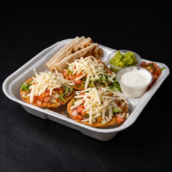 Mexican food platter with cheese-topped tostadas, folded tacos, fresh vegetables, guacamole, and sour cream in a takeout tray.