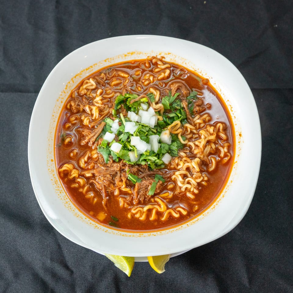 Bowl of spicy red noodle soup topped with shredded meat, cilantro, diced onions, and lime wedges.