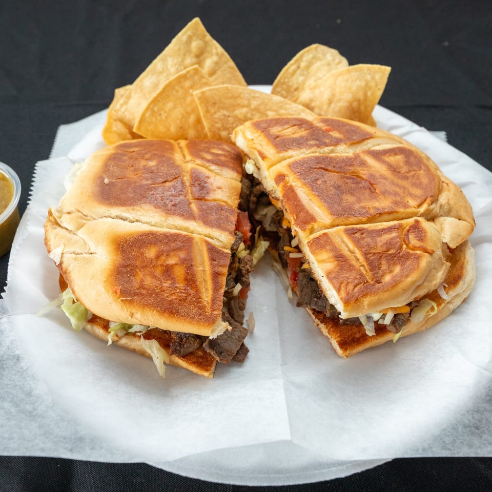 Two toasted steak sandwiches with lettuce and cheese, served with tortilla chips and green and red dipping sauces.