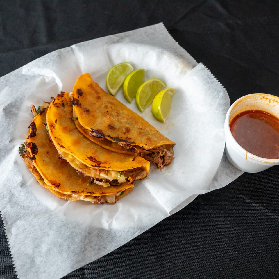 Three crispy birria tacos filled with shredded meat, served with lime wedges and a cup of consommé on parchment paper.
